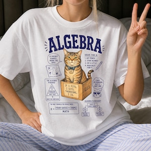 May include: A white t-shirt featuring a graphic of a tabby cat sitting in a cardboard box. The design includes the text ALGEBRA, maths-themed diagrams, a flowchart, and humorous captions about solving for X, including the phrase IF I FITS, I SITS.