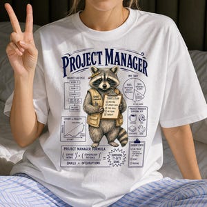May include: A white t-shirt featuring a graphic of a raccoon wearing a tan vest and holding a clipboard labelled Chaos Coordinator. The design includes text reading Project Manager, a project life cycle chart, a pie chart, and a formula.