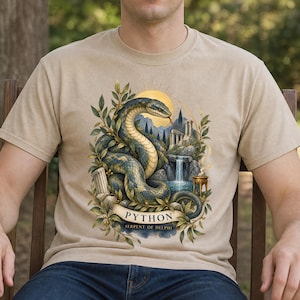 Könnte beinhalten: Ein beiges T-Shirt mit einer grafischen Illustration einer großen, gewundenen Schlange, umgeben von Lorbeerzweigen, einem Wasserfall, antiken Steinsäulen und einer goldenen Sonne. Ein Banner zeigt den Text PYTHON SERPENT OF DELPHI.