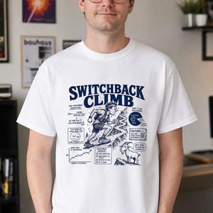Puede incluir: Una camiseta blanca de manga corta con un estampado gráfico azul oscuro titulado SWITCHBACK CLIMB. El diseño incluye un corredor en un sendero sinuoso, una cabra, varios gráficos y texto como The Switchback Equation y Switchback Checklist.