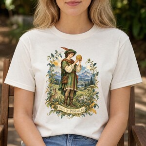 May include: A cream-colored t-shirt featuring a graphic illustration of a figure in a green tunic and hat holding a golden coin. The design includes a rabbit, mountains, and foliage, with a banner at the bottom reading Page of Pentacles.