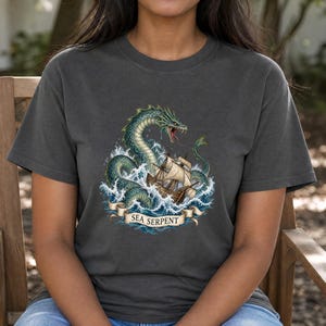 May include: A charcoal grey short-sleeve t-shirt featuring a graphic print of a green sea serpent emerging from ocean waves to attack a wooden sailing ship. Below the illustration, a beige banner displays the text SEA SERPENT in black letters.
