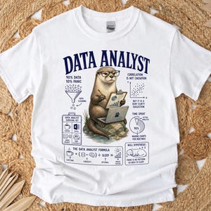 Op de afbeelding: Een wit t-shirt met een afbeelding van een otter met een bril, die een klembord en een laptop vasthoudt. De tekst luidt DATA ANALYST, 90% DATA 10% PANIC, DATA CLEANING, CORRELATION IS NOT CAUSATION, TIME SPENT en THE DATA ANALYST FORMULA.