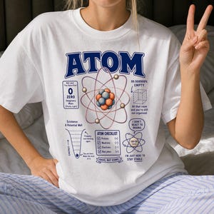 May include: A white t-shirt featuring a large blue graphic of an atom with scientific text including "ATOM," "0 ZERO Imaginary Mass," "99.999999% EMPTY," "ATOM CHECKLIST," "Protons (+)," "Neutrons (-)," "Electrons (-)," and "Bad jokes (✓)."