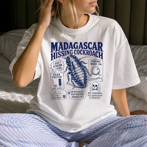 Pode incluir: Uma t-shirt branca com um gráfico azul de uma barata-bufadora de Madagáscar. O design inclui texto como MADAGASCAR HISSING COCKROACH, I DON'T SCREAM. I HISS., HISSING MODE: ON, FEAR FACTOR e outras etiquetas que detalham os traços do inseto.