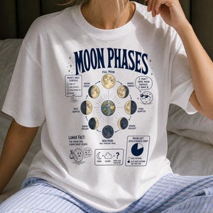 Puede incluir: Una camiseta blanca con un gráfico azul oscuro titulado MOON PHASES. El diseño incluye un diagrama circular de los ciclos lunares alrededor de una ilustración de la Tierra, acompañado de textos humorísticos, un horario lunar y un gráfico de eficacia.