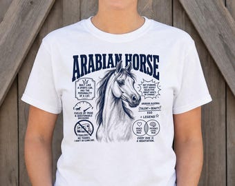 Puede incluir: Una camiseta blanca de manga corta con un gráfico azul oscuro de una cabeza de caballo árabe rodeada de texto e iconos, incluyendo frases como Built like a sports car, Arabian Algebra y Every ride is a negotiation.