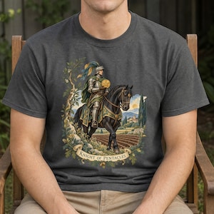 May include: A charcoal grey t-shirt featuring a graphic print of a knight in silver armour riding a black horse. The knight holds a gold pentacle. The scene includes a landscape with fields, trees, and a banner with the text Knight of Pentacles.