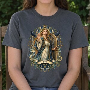 May include: A charcoal gray t-shirt featuring a graphic illustration of a woman with long blonde hair wearing a blue and white robe, holding one hand up in a stop gesture. The design includes two black ravens, golden scales, and the text Syn.