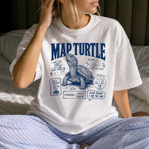 May include: A white short-sleeve t-shirt featuring a blue graphic of a map turtle. The design includes the text "MAP TURTLE" and various diagrams labeled "I DON'T GET LOST," "MY GPS," "SHELL TERMINAL," "A DAY IN MY LIFE," and "SLOW DOWN? I AM THE MAP."