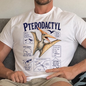 Puede incluir: Una camiseta blanca con una ilustración grande y detallada de un Pterodactyl en el centro. El gráfico incluye texto y diagramas en azul, como una lista de tareas, un medidor de carga alar y detalles esqueléticos, con el título PTERODACTYL arriba.