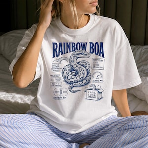 Puede incluir: Una camiseta blanca de manga corta con un estampado gráfico azul de una serpiente enroscada. El texto dice RAINBOW BOA, TODAY'S PLAN: EAT, SHED, JUDGE YOU, SASS ALWAYS MAX, e IF I FITS, I SITS & AMBUSHES, junto con varios diagramas científicos.