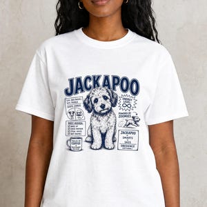 May include: A white t-shirt featuring a dark blue graphic of a Jackapoo dog. The design includes the text JACKAPOO, a daily agenda list, energy level details, and a small illustration of a running dog with the caption powered by zoomies.