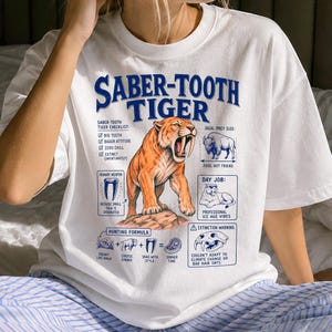 Puede incluir: Una camiseta blanca de manga corta con un gráfico azul y naranja de un tigre dientes de sable. El diseño incluye texto como Saber-Tooth Tiger Checklist, Ideal Prey Size, Day Job y una fórmula de caza con ilustraciones de un cráneo.