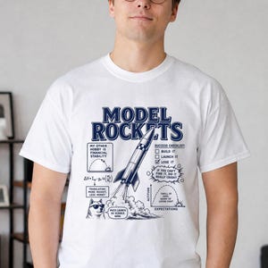 May include: A white short-sleeved t-shirt featuring a dark blue graphic of a rocket with the text "MODEL ROCKETS." The design includes a success checklist, a physics equation, a graph of altitude versus expectations, and a dog wearing sunglasses.