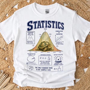May include: A white t-shirt featuring a graphic print titled STATISTICS. The design includes a bell curve with a turtle in the centre, various mathematical formulas, a scatter plot, a pie chart, and text including "AVERAGE? MORE LIKE AWFUL-AGE."