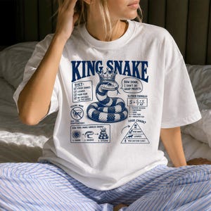 May include: A white short-sleeved t-shirt featuring a blue graphic print of a snake wearing a crown. The design includes text such as KING SNAKE, diet lists, a slither formula, and a food chain pyramid, all rendered in a detailed, illustrative style.