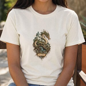 以下が含まれることがあります： クリーム色の半袖Tシャツ。木々や小さな滝のある岩場に巻き付いた、長い蛇のようなドラゴンの詳細なグラフィックプリントが施されています。イラストの基部にあるバナーには「DILONG」という文字が印刷されています。