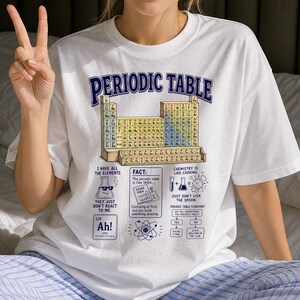 May include: A white t-shirt featuring a blue graphic of the periodic table of elements. The design includes text such as Periodic Table, I have all the elements, Chemistry is like cooking, and a flowchart labelled Periodic Table Flowchart.