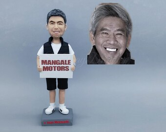 May include: A custom bobblehead figurine of a man holding a sign that reads "Mangale Motors." The figurine is wearing a white shirt, black vest, and shorts. A grayscale photo of a smiling man is in the background.