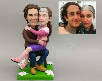 May include: A custom-made figurine of a couple, a man and a woman, sitting together. The man is wearing a brown jacket and blue jeans, and the woman is wearing a pink jacket and purple shorts. The figurine is made of clay and is painted in realistic colours.