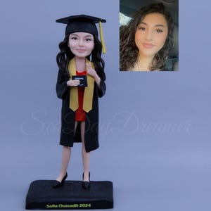 Puede incluir: Una figura de graduación de una persona que lleva una toga y un birrete negros, sosteniendo un diploma. La figura está de pie sobre una base negra con el texto "Sofia Ouassadit 2024".