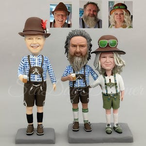 Op de afbeelding: Drie op maat gemaakte beeldjes van mensen in traditionele Duitse kleding. De beeldjes zijn gemaakt van klei en zijn geschilderd in realistische kleuren. De beeldjes houden bierkruiken vast en staan op een houten voet.