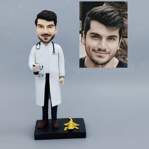 May include: A figurine of a doctor in a white coat, holding a game controller. The figurine has a stethoscope and is standing on a black base with a yellow frog. A photo of a man is in the background.