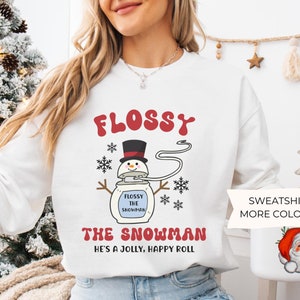 May include: White sweatshirt with the text "FLOSSY THE SNOWMAN" in red and black. The design features a snowman with a top hat and snowflakes. The phrase "HE'S A JOLLY, HAPPY ROLL" is also printed on the shirt.