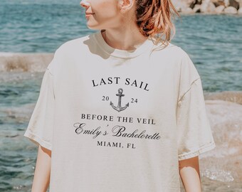 May include: A white t-shirt with the text "Last Sail" and an anchor graphic. The text "Before the Veil" and "Emily's Bachelorette" are also printed on the shirt. The shirt is worn by a person standing in front of a body of water.