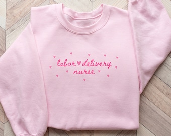 Könnte beinhalten: Rosafarbener Pullover mit dem Text "labor ♥ delivery nurse" in rosa Schrift, umgeben von kleinen rosa Herzen.
