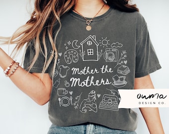 May include: A grey t-shirt with a white graphic design featuring a house, a crescent moon, a sun, a baby bottle, a baby onesie, a horseshoe, a washing machine, a cup of coffee, a fork and spoon, a bed, and a woman breastfeeding a baby. The text "Mother the Mothers" is written in white on the shirt. The text "Ourma Design Co." is written in white on a white banner in the bottom right corner of the image.