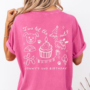 以下が含まれることがあります： 背面に白いグラフィックデザインが施されたピンク色のTシャツ。デザインには犬、カップケーキ、パーティーハット、「I wa let the dogs out?」と「Jonny's 2nd Birthday」の文字が含まれています。