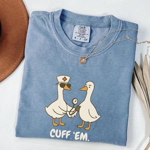 May include: A blue Comfort Colors t-shirt featuring a graphic of two cartoon geese. One goose wears a nurse's hat and sunglasses, holding a blood pressure cuff. The text "CUFF 'EM" is printed below the geese. A gold necklace is draped on the shirt.