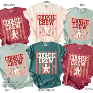 May include: Six short-sleeved t-shirts in crimson, ivory, bay, and blue spruce colors. Each shirt features a graphic with vertical stripes, the text "COOKIE CREW" in bold letters, and a central illustration of a gingerbread person holding a rolling pin.