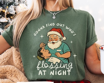 May include: Green t-shirt with a graphic of Santa Claus as a dentist, examining a patient. The text reads "Gonna find out who's flossing at night." The shirt is a comfortable, casual style.