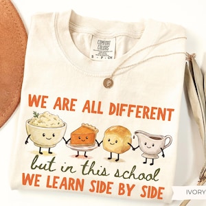 May include: An ivory t-shirt featuring a graphic of four anthropomorphic food items holding hands. The text reads, "WE ARE ALL DIFFERENT but in this school WE LEARN SIDE BY SIDE." The food items include a bowl of mashed potatoes, a slice of pumpkin pie, a dinner roll, and a gravy boat.