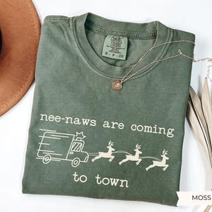 May include: A moss green short-sleeve t-shirt with a crew neckline and a Comfort Colors label. The graphic features a white line drawing of an emergency vehicle and three reindeer, with the text "nee-naws are coming to town" printed in white.