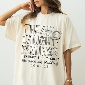 May include: Cream-colored t-shirt with the text "THEY CAUGHT FEELINGS" in a bold, outlined font. Below, it reads "I CAUGHT THIS T-SHIRT the jackson WEDDING 10.05.25". The design includes a ring box and disco ball illustration.