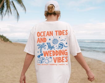 May include: White t-shirt with the text "OCEAN TIDES AND WEDDING VIBES" in orange and blue. The design includes illustrations of a palm tree, beach chair, disco ball, wave, sunglasses, and rings. The text "The Jackson wedding | Destin, FL" is also printed on the shirt.