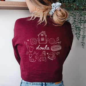 May include: A maroon sweatshirt with a white graphic design of a house, a baby, a stethoscope, a heart, a horseshoe, a yoga ball, a rolled up towel, and the word "doula".