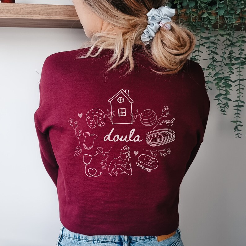 May include: A maroon sweatshirt with a white graphic design of a house, a baby, a stethoscope, a heart, a horseshoe, a yoga ball, a rolled up towel, and the word "doula".