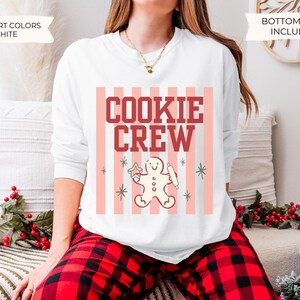 May include: A white long-sleeve crewneck sweatshirt featuring a graphic print with vertical light pink stripes, the text "COOKIE CREW" in dark red block letters, and a central illustration of a gingerbread person holding a rolling pin.