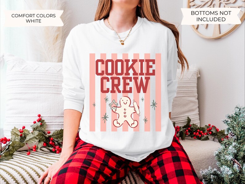 May include: A white long-sleeve crewneck sweatshirt featuring a graphic print with vertical light pink stripes, the text "COOKIE CREW" in dark red block letters, and a central illustration of a gingerbread person holding a rolling pin.