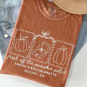 May include: Rust-colored t-shirt with a white graphic design featuring pumpkins and a diamond ring in a box. The text "fresh off the pumpkin patch" and "Sarah's Bachelorette Ellijay, GA" are also printed on the shirt. The shirt is paired with blue jeans and a necklace.