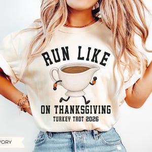 May include: An ivory short-sleeve t-shirt featuring a graphic of a gravy boat with arms and legs running. The text reads RUN LIKE ON THANKSGIVING TURKEY TROT 2026 in black block lettering. The gravy boat is white with brown liquid inside.
