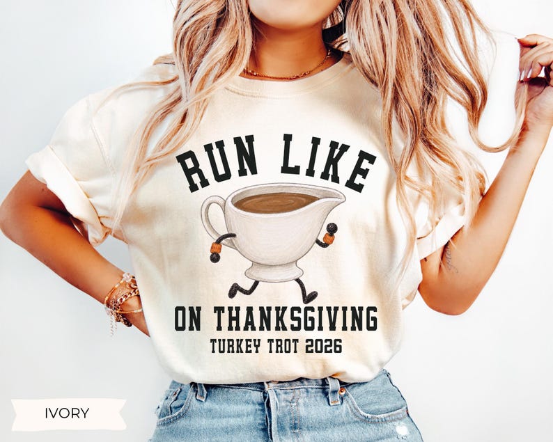 May include: An ivory short-sleeve t-shirt featuring a graphic of a gravy boat with arms and legs running. The text reads RUN LIKE ON THANKSGIVING TURKEY TROT 2026 in black block lettering. The gravy boat is white with brown liquid inside.