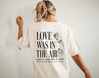 May include: Cream-colored t-shirt with the text "LOVE WAS IN THE AIR" in a bold, black font. Below the text is "and so was this t-shirt" and the names "JUSTIN AND ERICA | 6.7.2025". A rose and ring graphic is on the right.