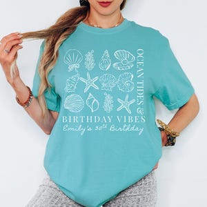 May include: A turquoise t-shirt with a white seashell and starfish design. The shirt has the text "Ocean Tides & Birthday Vibes" and "Emily's 30th Birthday" printed on it. The shirt is short-sleeved.