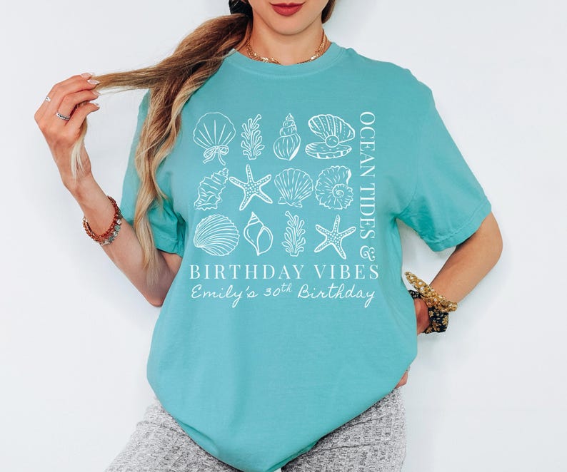 May include: A turquoise t-shirt with a white seashell and starfish design. The shirt has the text "Ocean Tides & Birthday Vibes" and "Emily's 30th Birthday" printed on it. The shirt is short-sleeved.